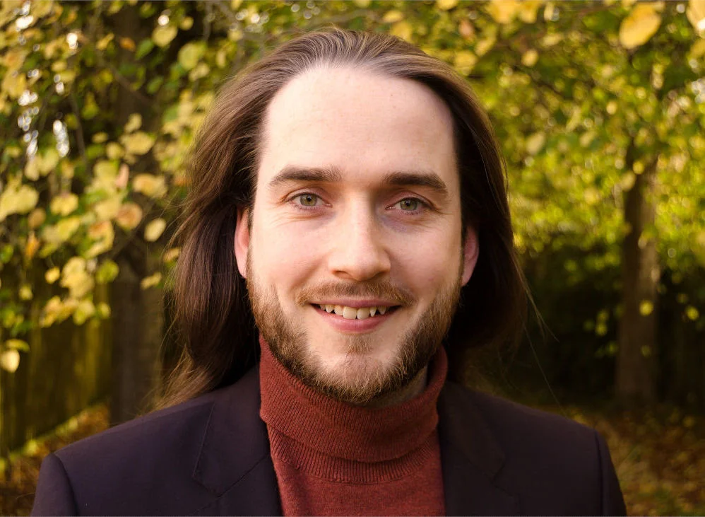 A headshot of Galen, who is a man with a short beard. He is wearing an orange roll-neck jumper and black suit jacket.