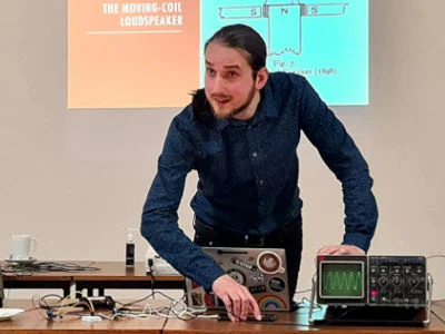 Galen leans over a laptop, oscilloscope, and wires on a table. A projection reads '1898 The Moving-Coil Loudspeaker'.