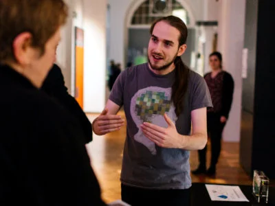 Galen gestures with both hands while speaking to a person on the left of the frame who is out-of-focus. 